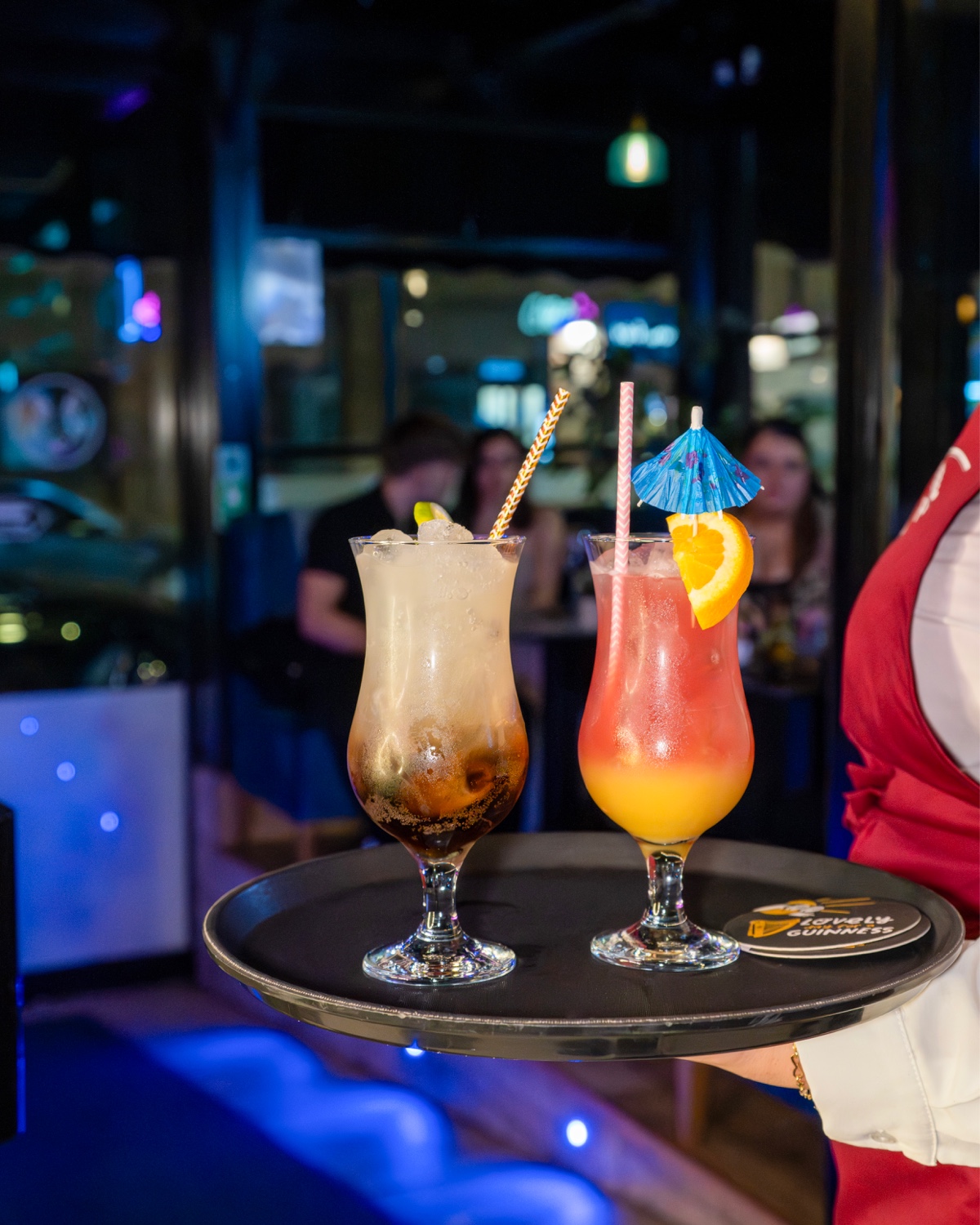 A waiter carries a tray with two long-glass cocktails through the bar